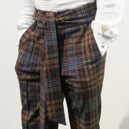 Boot-cut pants featuring a brown and black checkered pattern, a high-waisted design with a large matching fabric waist tie, and a flared leg opening.