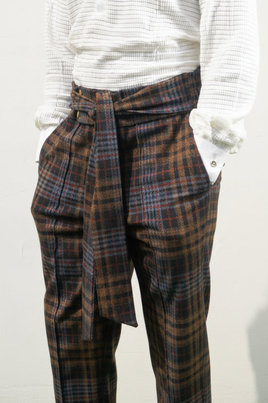 Boot-cut pants featuring a brown and black checkered pattern, a high-waisted design with a large matching fabric waist tie, and a flared leg opening.