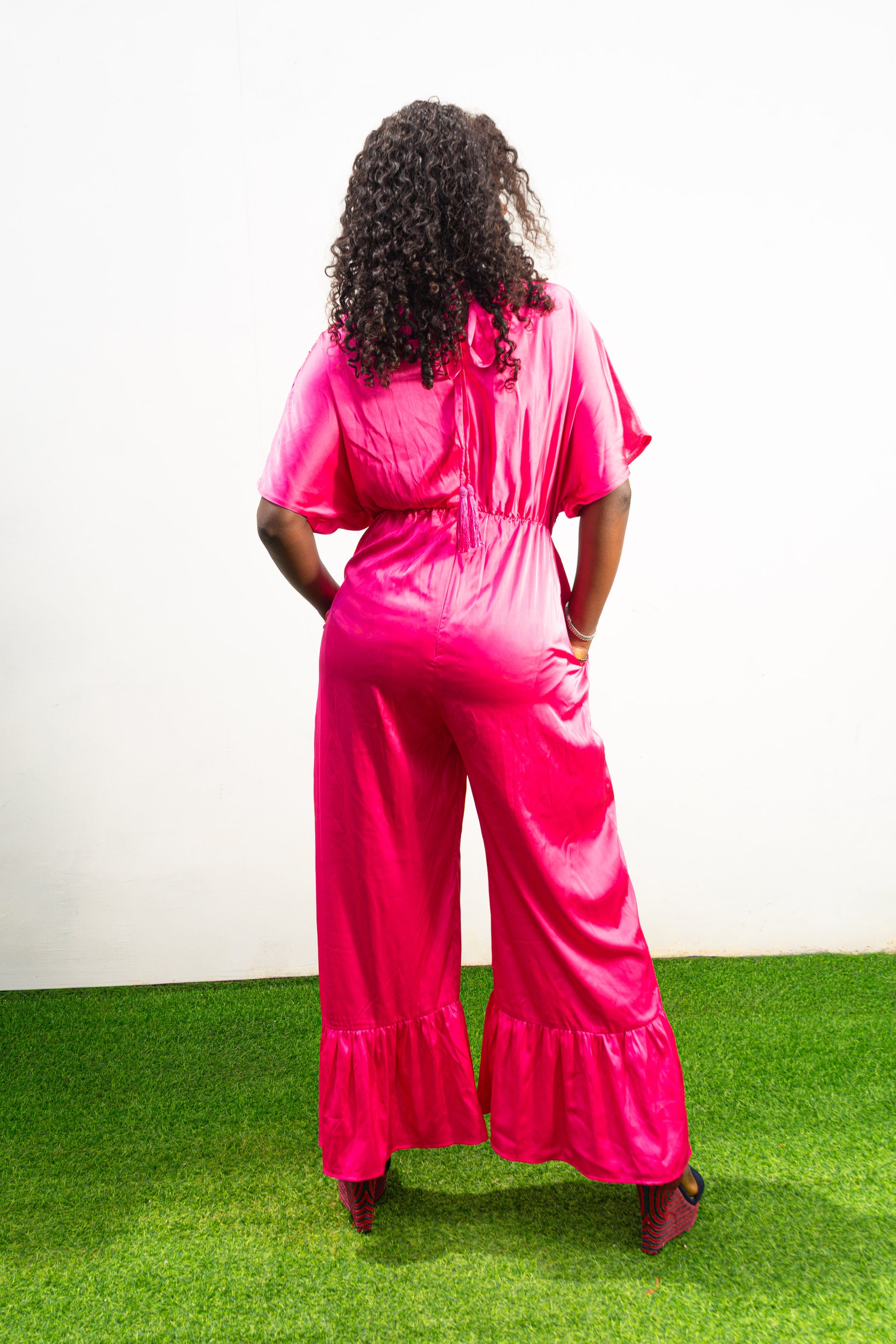 Smocked Pink Jumpsuit featuring a vibrant fuchsia fabric with a beaded V-shape design on the bodice, short flutter sleeves, and ruffled ankle hems. Back View