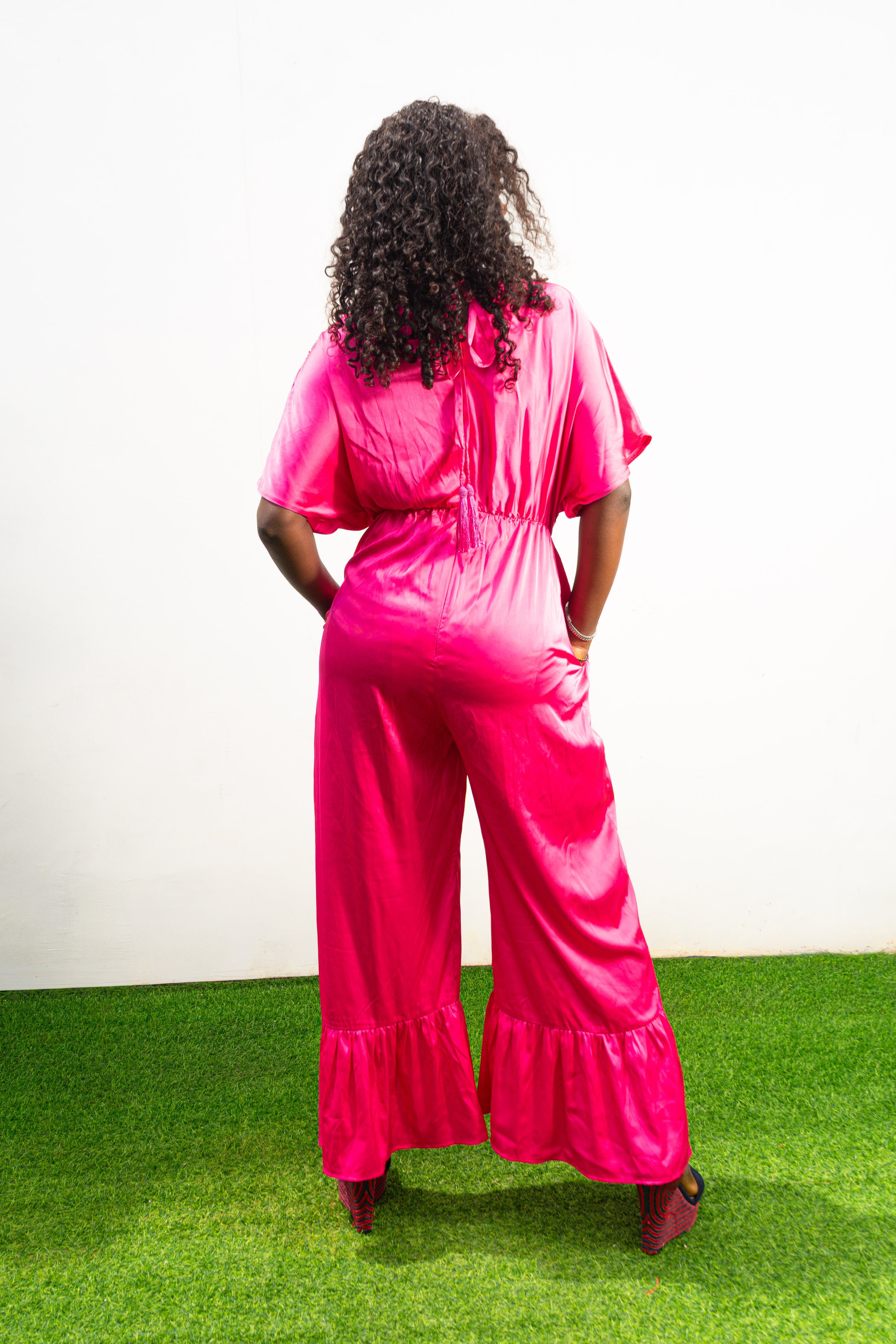 Smocked Pink Jumpsuit featuring a vibrant fuchsia fabric with a beaded V-shape design on the bodice, short flutter sleeves, and ruffled ankle hems. Back View