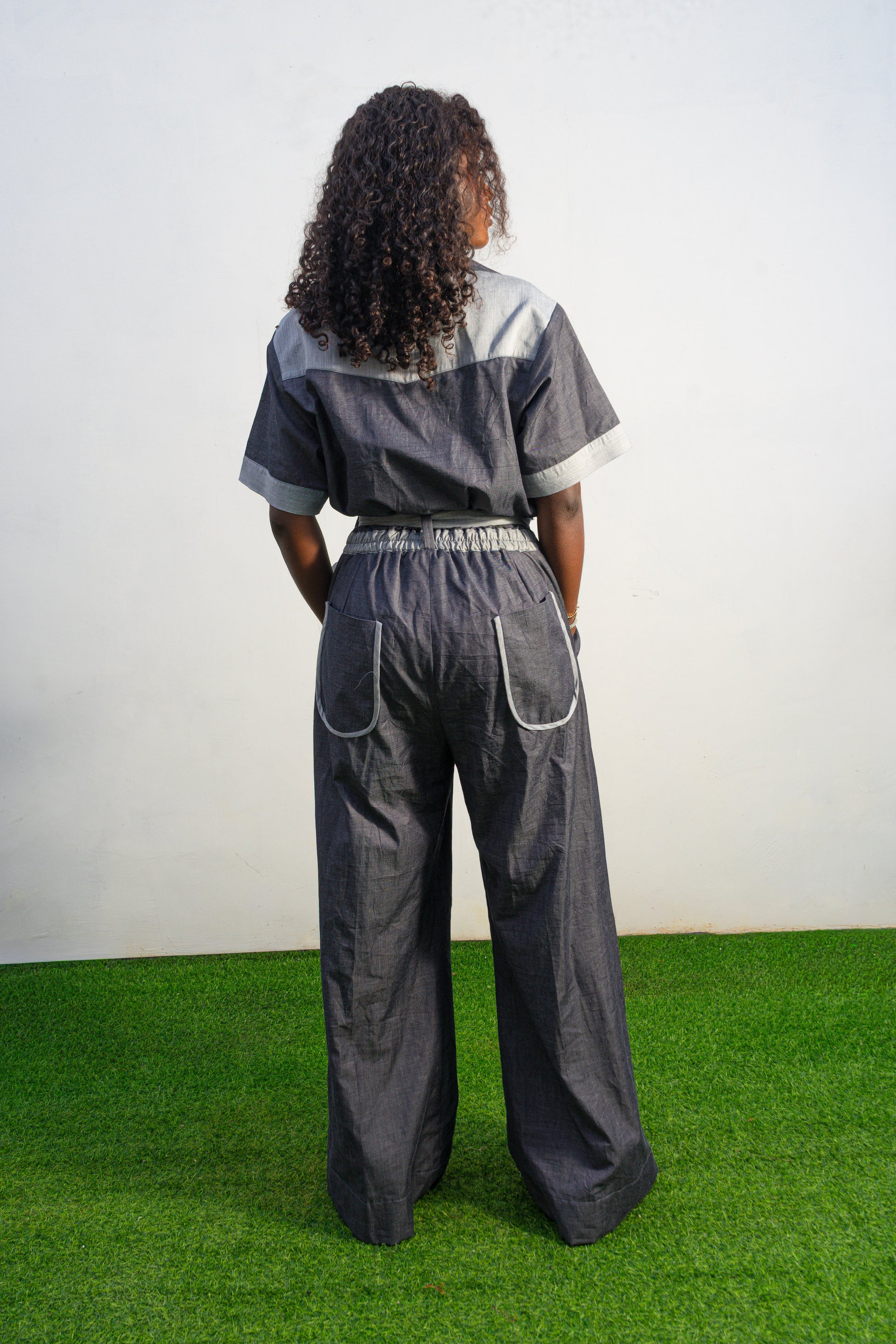 Trendy Denim Jean Jumpsuit featuring a deep indigo denim fabric, light-wash contrast trim on the short sleeves and pockets, and a matching light-wash denim waist tie. Back View