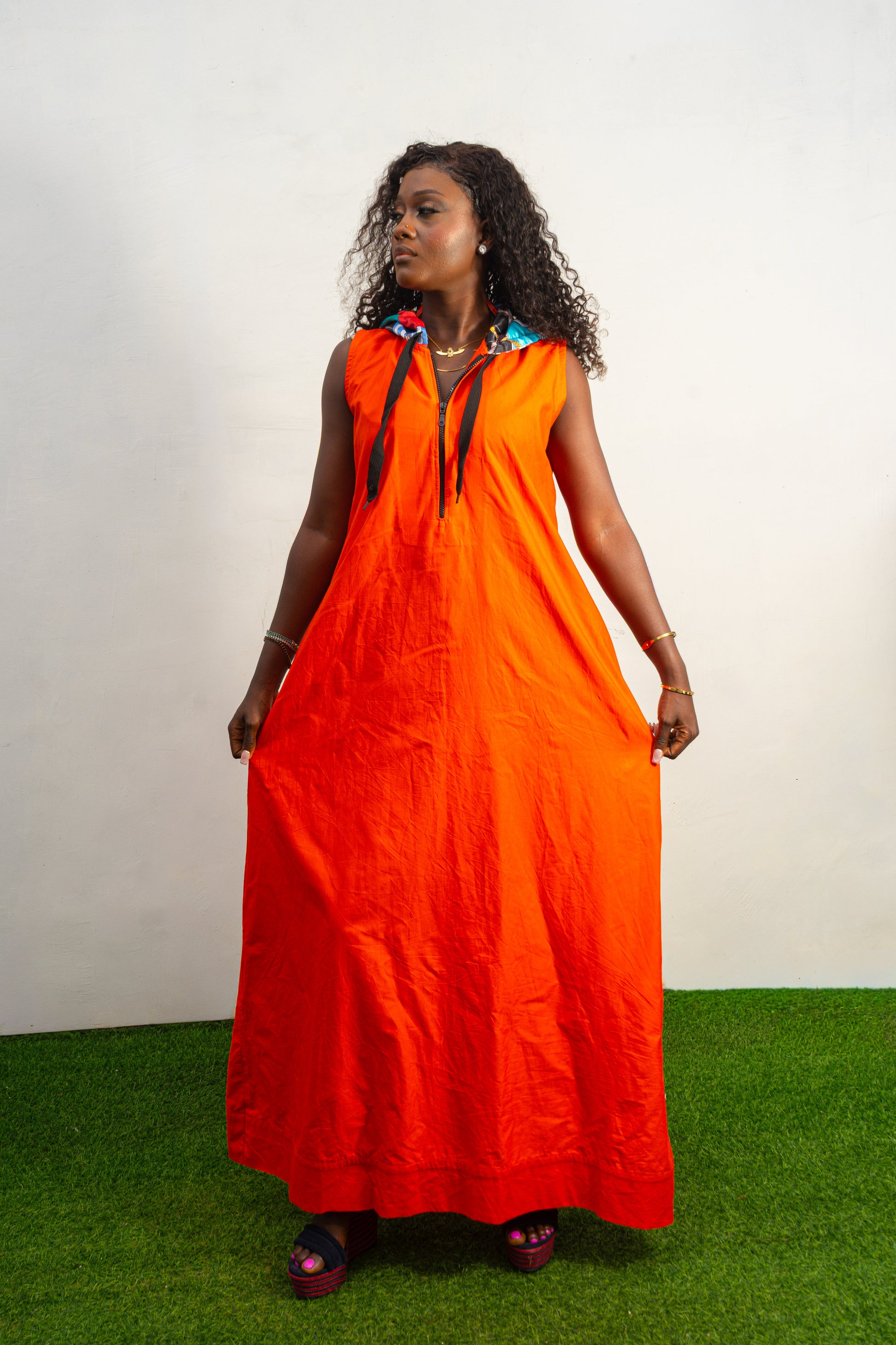 Jumpsuit featuring a vibrant solid orange hue, a deep V-neckline with a metal zipper and black pull cords, and a modern hoodie lined with an intricate, multi-colored Afrocentric pattern.