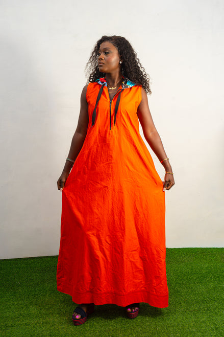 Jumpsuit featuring a vibrant solid orange hue, a deep V-neckline with a metal zipper and black pull cords, and a modern hoodie lined with an intricate, multi-colored Afrocentric pattern.