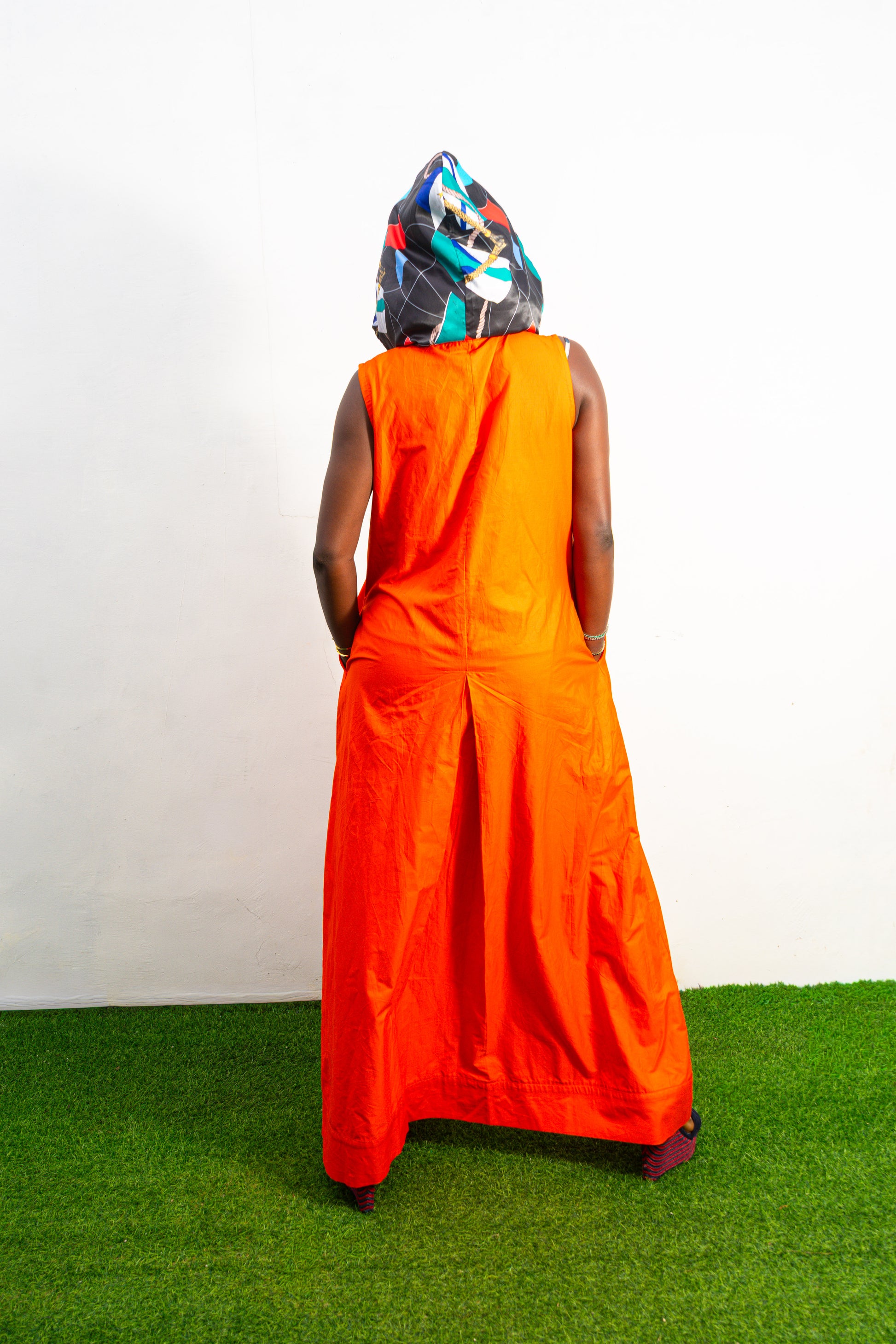 Jumpsuit featuring a vibrant solid orange hue, a deep V-neckline with a metal zipper and black pull cords, and a modern hoodie lined with an intricate, multi-colored Afrocentric pattern.Back View