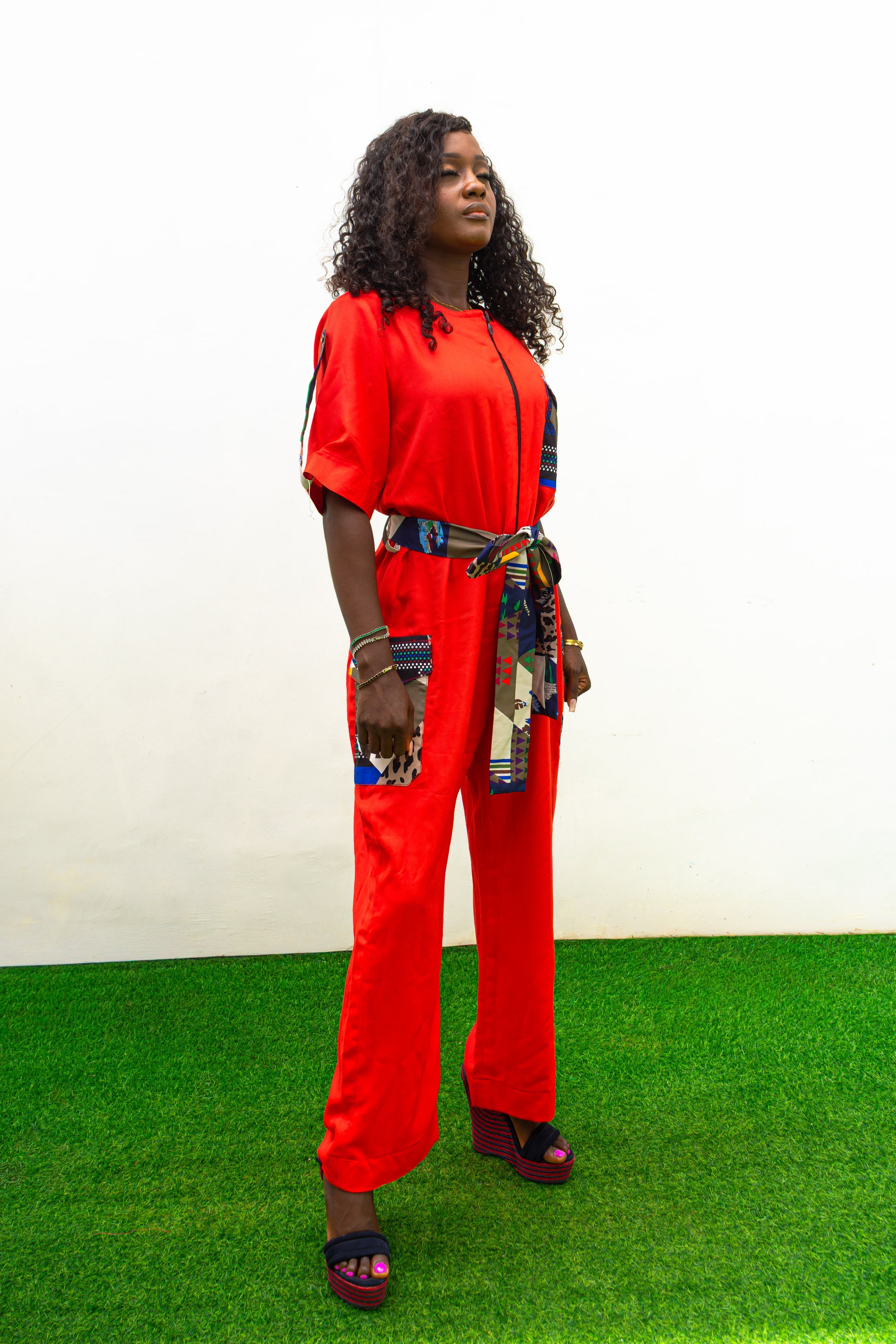 Chic Orange Jumpsuit with a colorful tassel  featuring a bright orange fabric, short sleeves, a patterned chest pocket, and a matching colorful patterned waist belt.