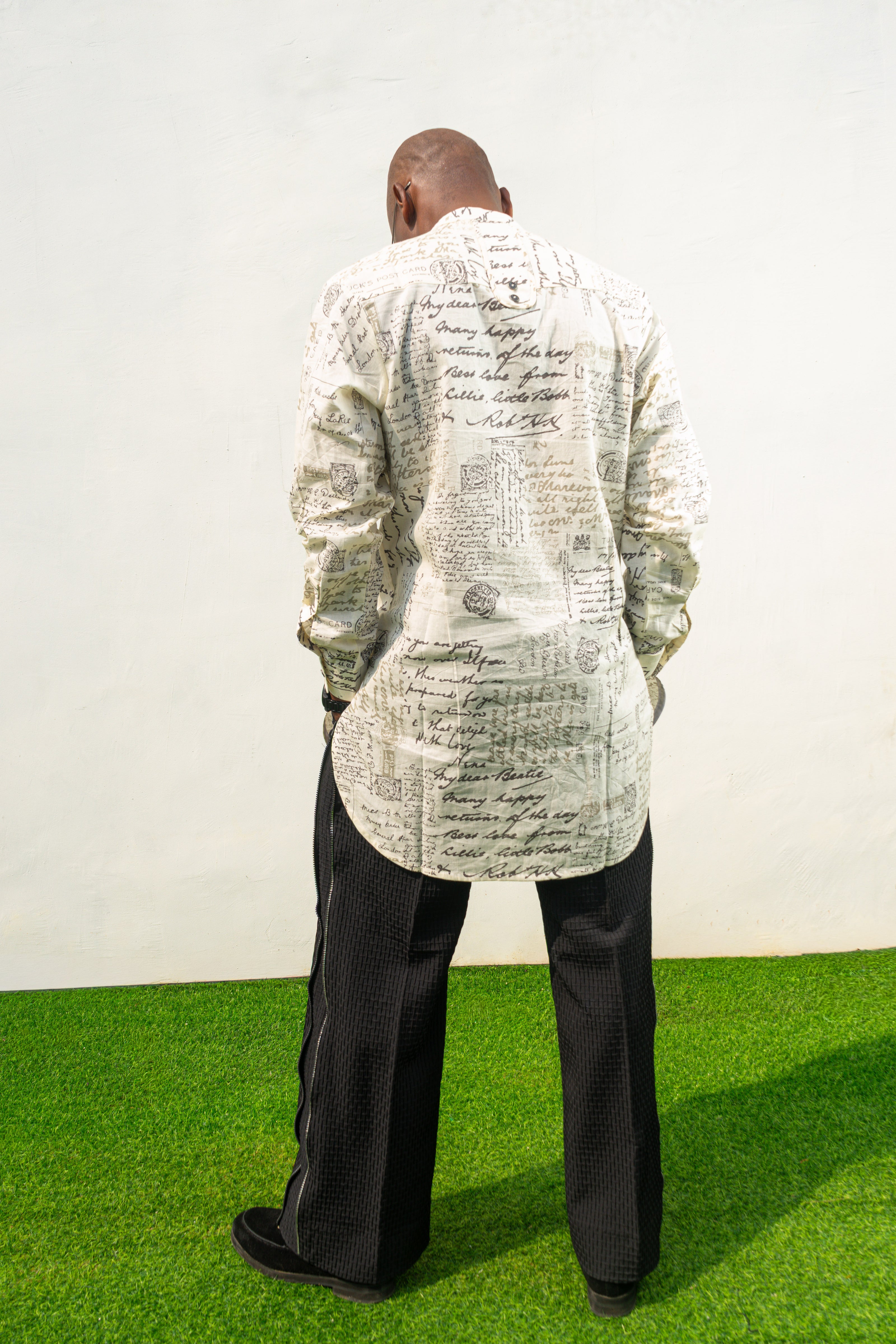 Linen Digital Shirt featuring a cream long-line tunic with black script patterns, a mandarin collar, and side slits paired with black textured wide-leg trousers. Back view.