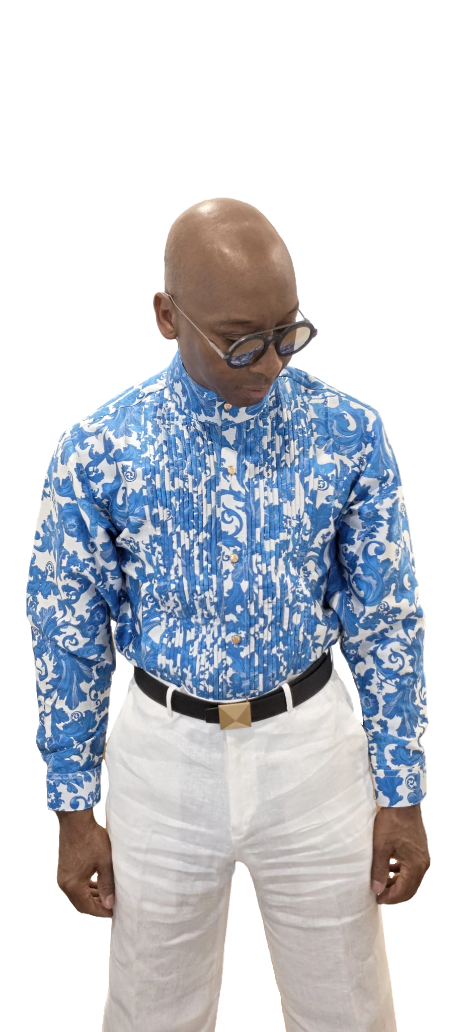 A man wearing a blue and white patterned long-sleeve button-up shirt, white trousers, and glasses, standing against a plain white background.