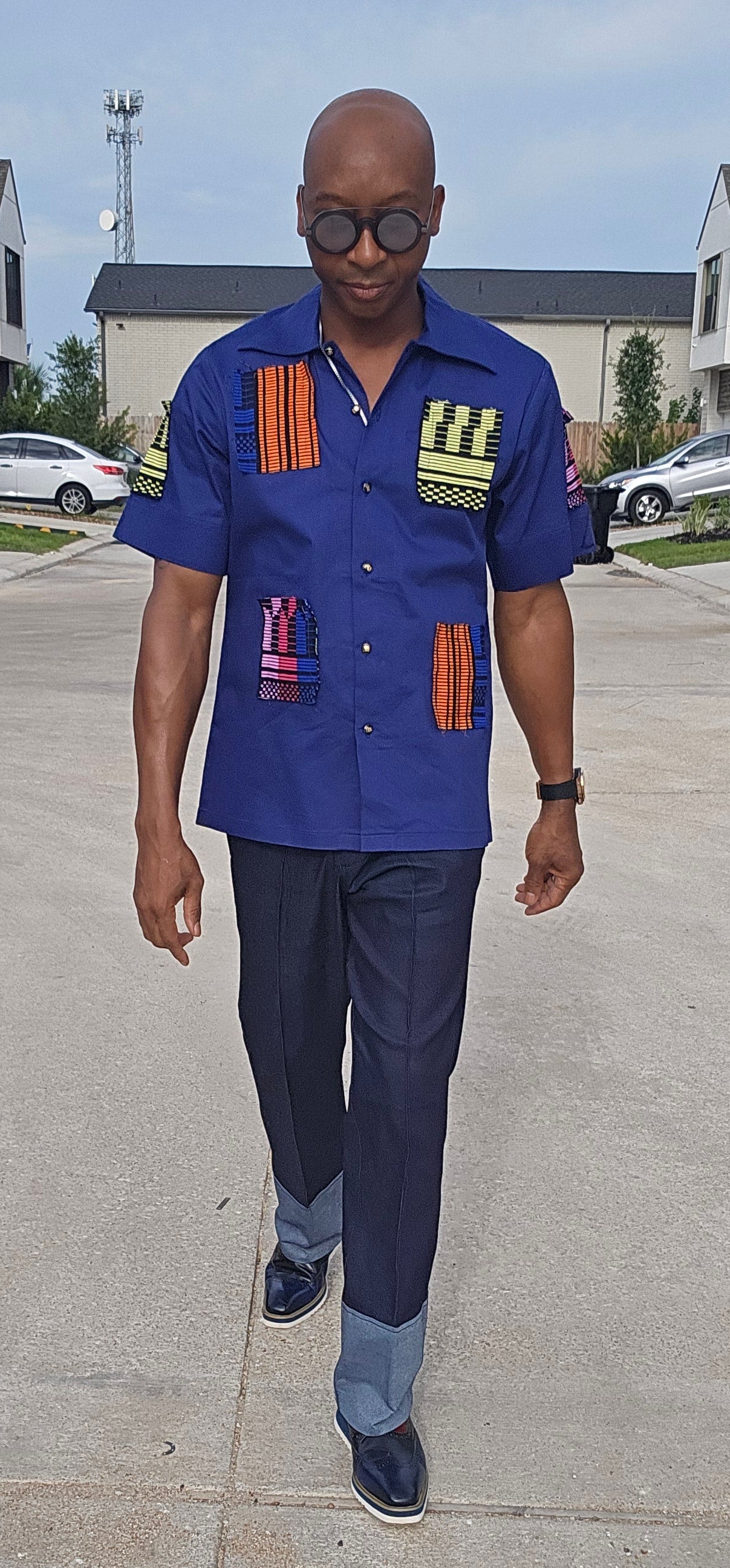 Shirt featuring a short-sleeved royal blue button-down design with vibrant geometric African print patches on the chest, sleeves, and front waist.