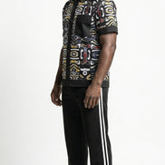 Shirt featuring a short-sleeved button-down silhouette dominated by an abstract, geometric Ankara pattern in white, yellow, and red tones, framed by a solid black collar, pocket, placket, and sleeve bands.