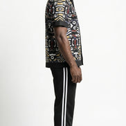Shirt featuring a short-sleeved button-down silhouette dominated by an abstract, geometric Ankara pattern in white, yellow, and red tones, framed by a solid black collar, pocket, placket, and sleeve bands.