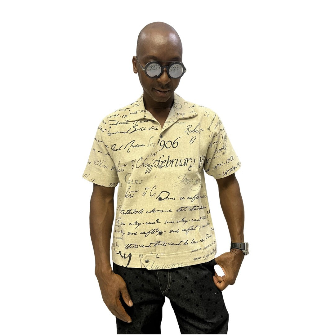 Short-sleeve cotton shirt featuring a black cursive script pattern on a cream-colored fabric with a button-down front and camp collar.