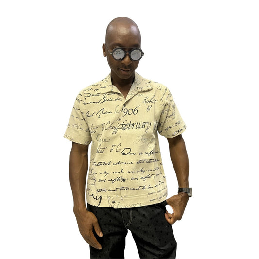 Short-sleeve cotton shirt featuring a black cursive script pattern on a cream-colored fabric with a button-down front and camp collar.