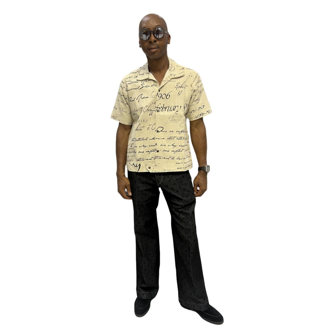 Short-sleeve cotton shirt featuring a black cursive script pattern on a cream-colored fabric with a button-down front and camp collar.
