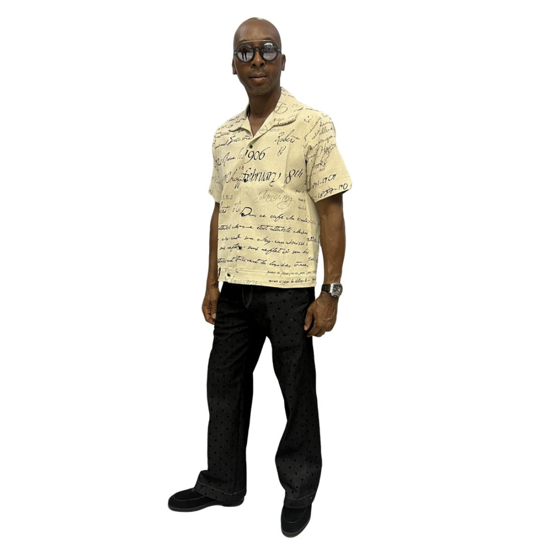 Short-sleeve cotton shirt featuring a black cursive script pattern on a cream-colored fabric with a button-down front and camp collar.
