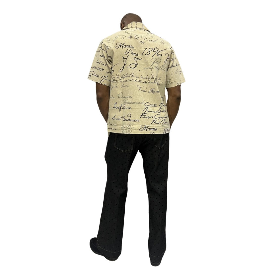 Short-sleeve cotton shirt featuring a black cursive script pattern on a cream-colored fabric with a button-down front and camp collar.