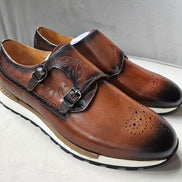 Double Monk Strap shoe featuring a hand-burnished tan leather upper with etched calligraphy, traditional medallion perforation on a rounded toe, and a contrasting white athletic sole.