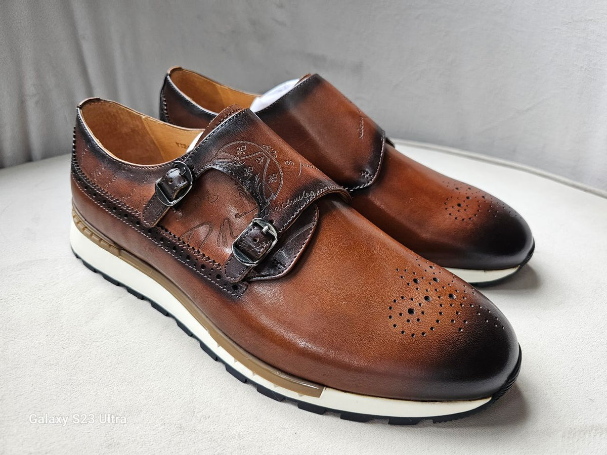 Double Monk Strap shoe featuring a hand-burnished tan leather upper with etched calligraphy, traditional medallion perforation on a rounded toe, and a contrasting white athletic sole.