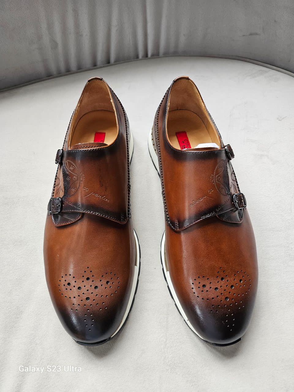 Double Monk Strap shoe featuring a hand-burnished tan leather upper with etched calligraphy, traditional medallion perforation on a rounded toe, and a contrasting white athletic sole.